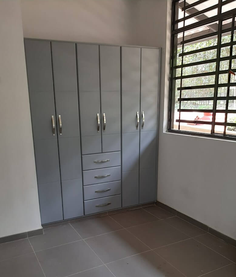 Clóset empotrado de diseño moderno en color gris claro, con puertas lisas verticales, cinco cajones centrales con tiradores curvos metálicos, ubicado junto a una ventana con reja negra en una habitación de piso cerámico gris.