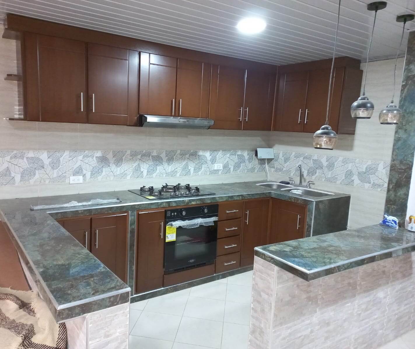 Cocina en forma de L con gabinetes de madera color caoba, mesón de mármol oscuro, estufa empotrada de cuatro puestos con horno, campana extractora y doble lavaplatos metálico.