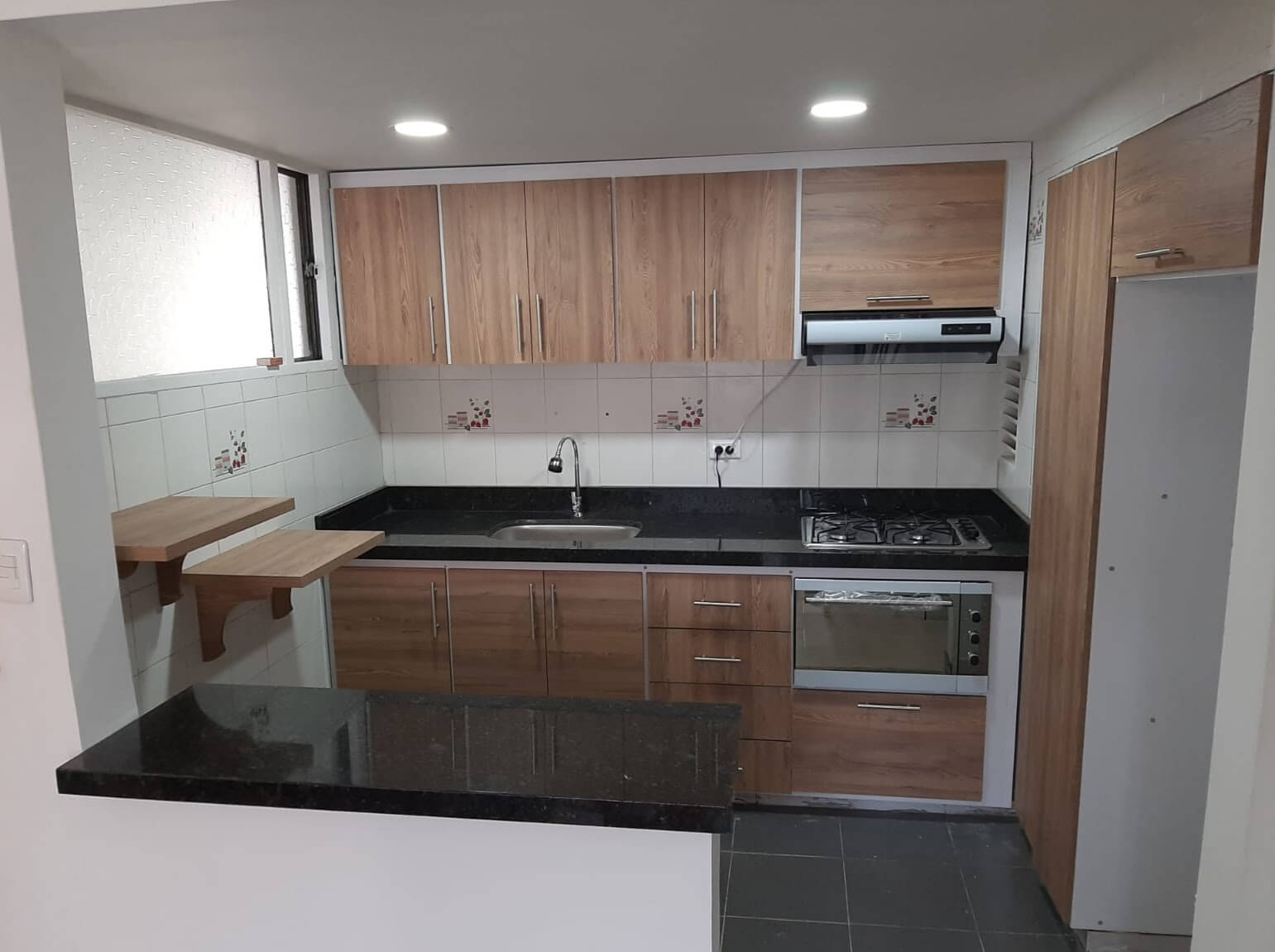 Cocina moderna con gabinetes de madera tono claro y marcos metálicos, mesón de granito negro, estufa empotrada de cuatro puestos con horno, campana extractora y lavaplatos sencillo.