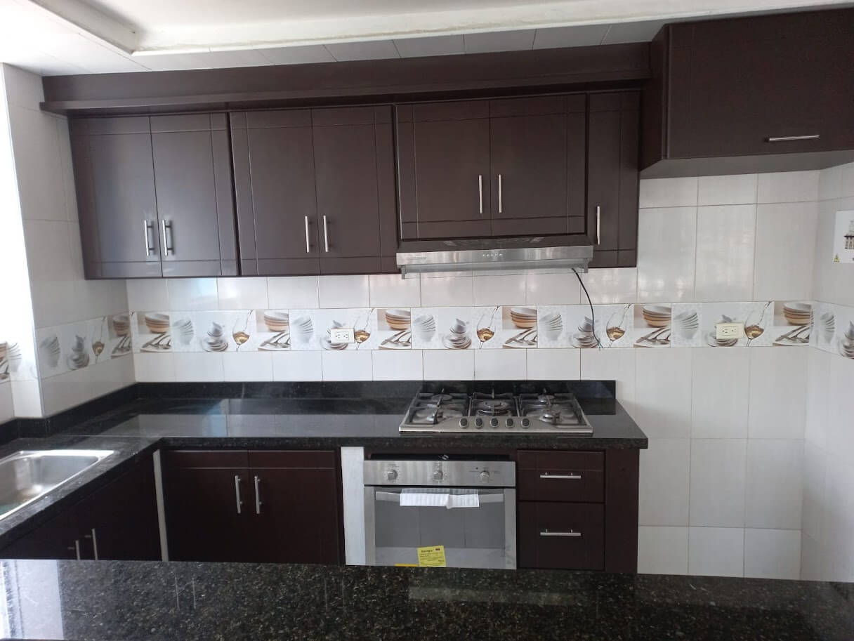 Cocina con gabinetes de madera tono wengué, mesón de granito negro, estufa de gas de cuatro puestos, horno empotrado, campana extractora y lavaplatos metálico doble.